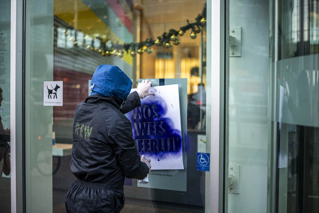 Rebel die met een sjabloon en krijtverf de tekst “NOS, Wees Eerlijk!” op een raam spuit.