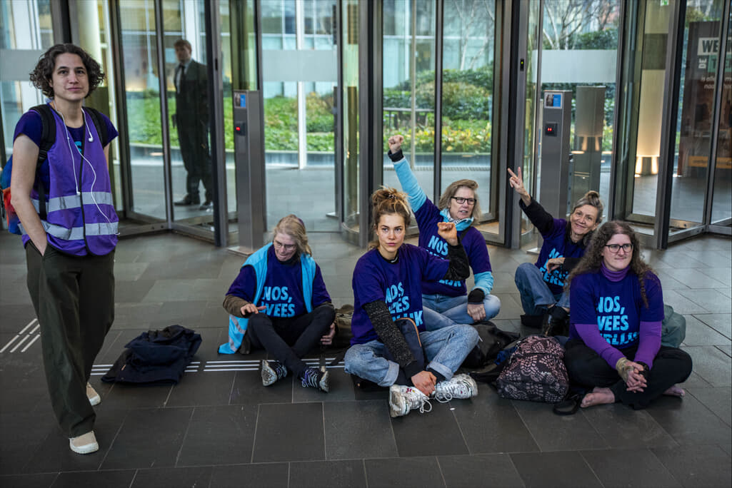 Groep rebellen die t-shirts met de tekst “NOS, Wees Eerlijk!” dragen en binnen op de grond zitten. Twee houden een vuist omhoog, één maakt een victory-sign. Daarnaast staat een rebel in een paars hesje.