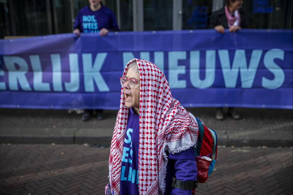 Roepende rebel met een keffiyeh over het hoofd met op de achtergrond een banner met de tekst “Meer Geld Voor Eerlijk Nieuws”.