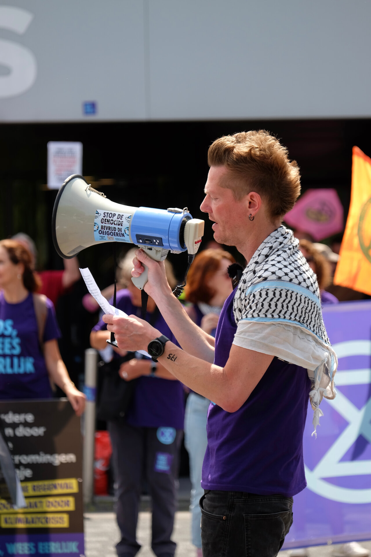 Martijn Dekker met megafoon in zijn rechterhand en A4 papier met tekst in zijn linkerhand. Hij draagt een paars t-shirt keffiyeh over zijn schouders. In de achtergrond zijn rebellen te zien