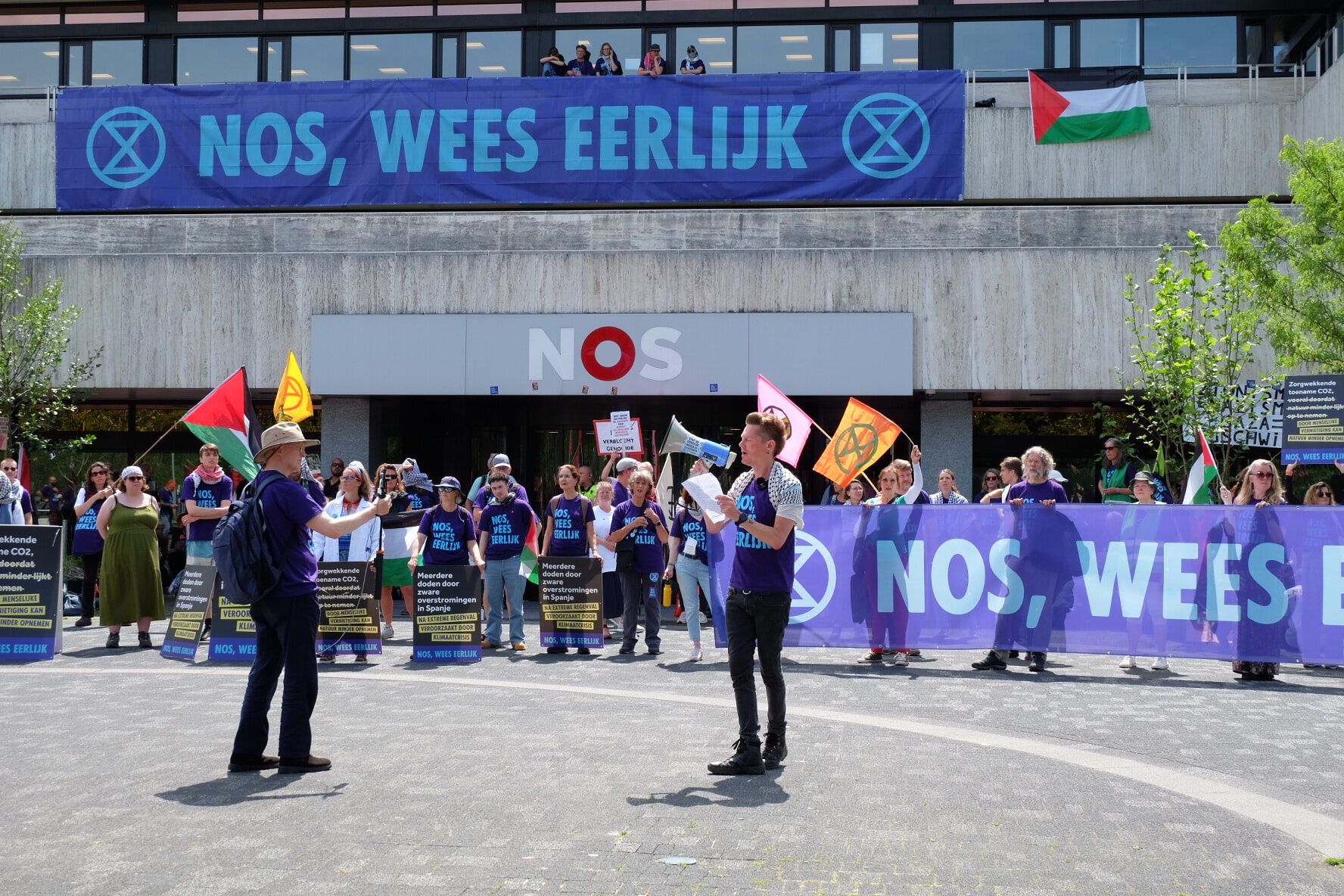 Martijn Dekker tijdens zijn speech met op de achtergrond het NOS-gebouw. Op het gebouw hangt een banner met de tekst “NOS, Wees Eerlijk” en XR-symbols aan weerszijde. Achter heb staan rebellen met protestborden en dezelfde banner. Een live-streamer die hem filmt staat links in beeld.