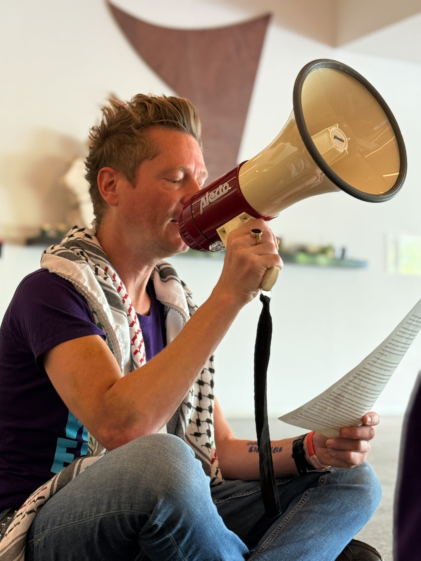 Martijn Dekker zittend op de grond in de lobby van het NOS-hoofdkantoor. Hij heeft een megafoon in zijn rechterhand en A4 papier met tekst in zijn linkerhand. Hij draagt een paars t-shirt en een keffiyeh over zijn schouders.