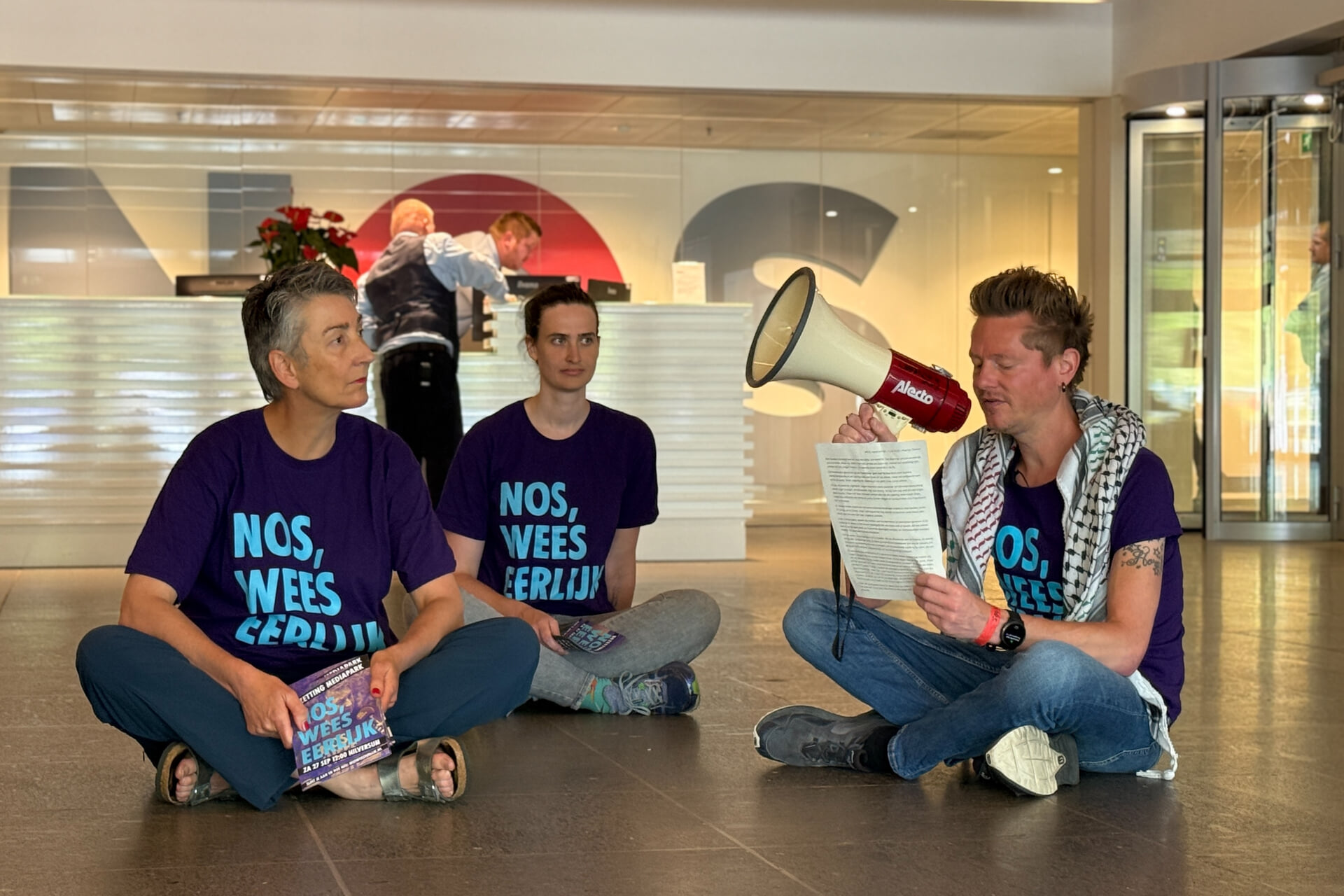 Martijn Dekker zit met twee andere rebellen op de grond in de lobby van het NOS-hoofdkantoor. Hij heeft een megafoon in zijn rechterhand en A4 papier met tekst in zijn linkerhand. Hij draagt een paars t-shirt met de tekst “NOS, Wees Eerlijk” en een keffiyeh over zijn schouders. In de achtergrond is de balie van de receptie te zien met enkele medewerkers.