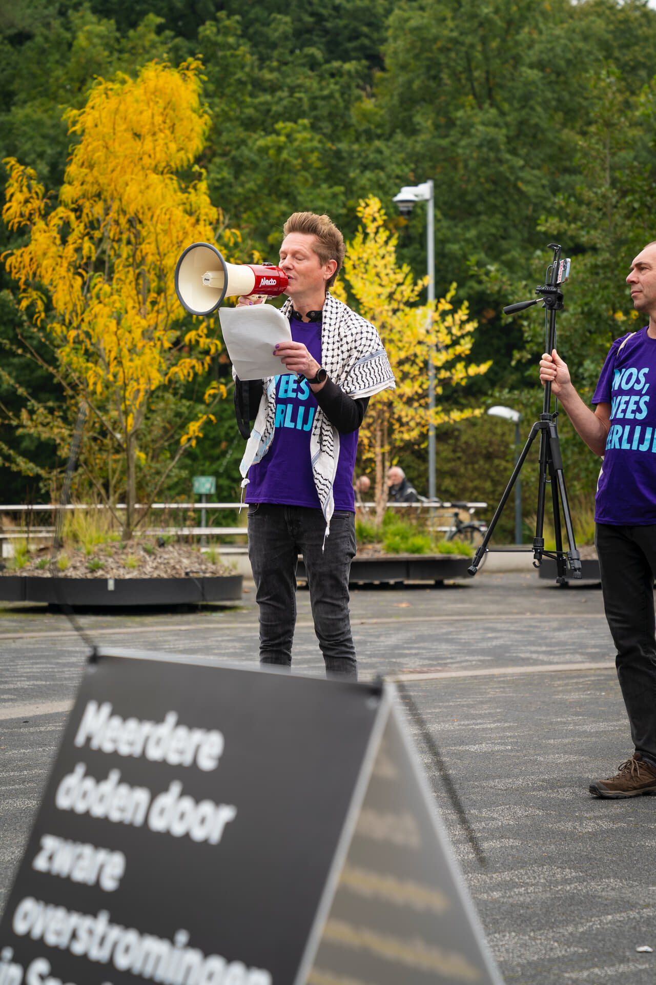 Martijn Dekker voor het NOS gebouw met megafoon in zijn rechterhand en A4 papier met tekst in zijn linkerhand. Hij draagt een paars t-shirt en een keffiyeh over zijn schouders. Links in beeld is een rebel te zien die aan het filmen is met een mobiele telefoon, vooraan in beeld een sandwichbord met een verbeterde kop van een NOS nieuwsartikel.